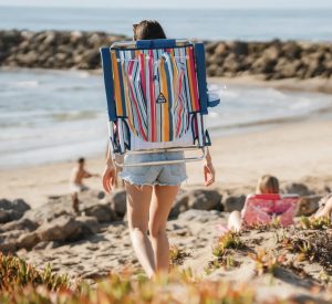 Person carrying a colorful striped GCI Outdoor Backpack Beach Chair on their back, walking toward the ocean on a sandy beach